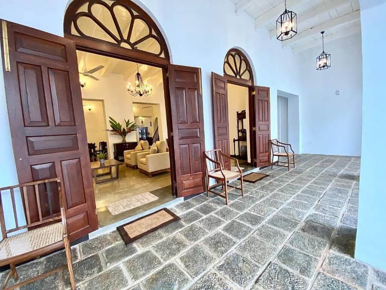 Spacious porch with stone flooring, graceful arches, wooden chairs, and open double doors leading to a well-lit living area with beige sofas and chandeliers—echoing the timeless elegance of Fort Galle.