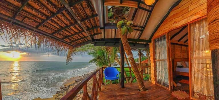 A wooden beach hut at Indika Beach Villa features an open veranda, a blue chair, and stunning ocean sunset views. Waves crash against rocks below, palm trees sway nearby—a tranquil Galle escape.