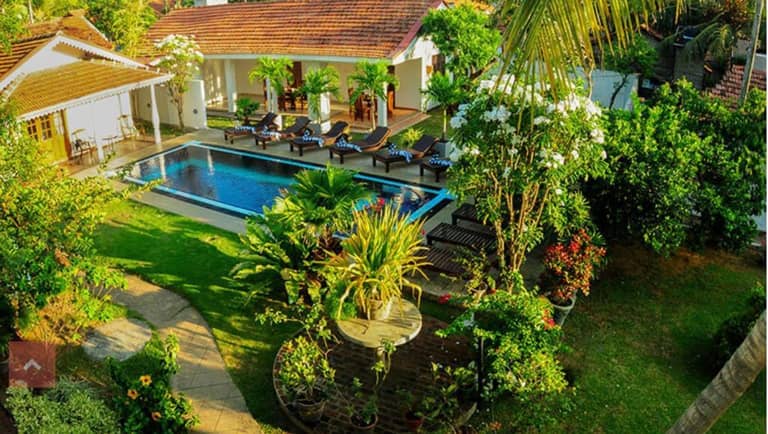 Aerial view of Sujeewani Villa in Negombo, featuring a tropical garden with a swimming pool, sun loungers, and two white buildings with red-tiled roofs, all surrounded by lush greenery.