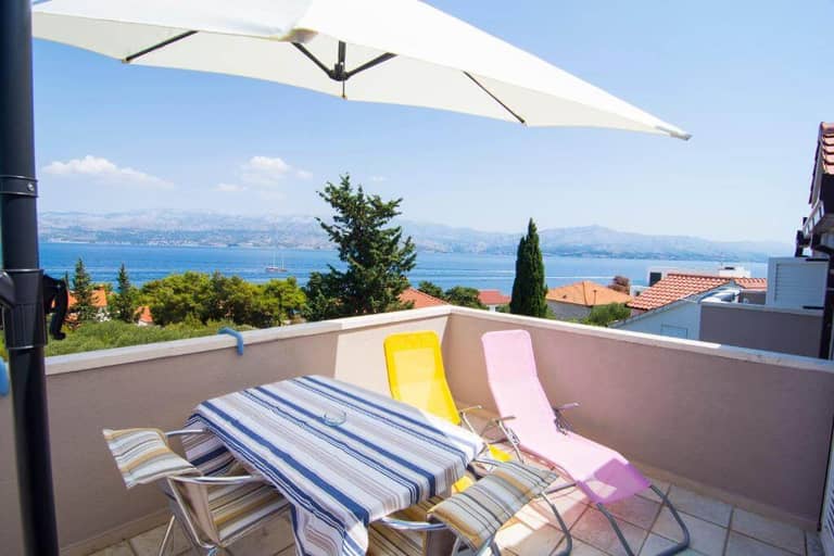 Outdoor terrace at Villa Tereza with a table, striped tablecloth, four chairs, two loungers, and a large umbrella, overlooking rooftops and greenery on Brac Island with mountain and water views.