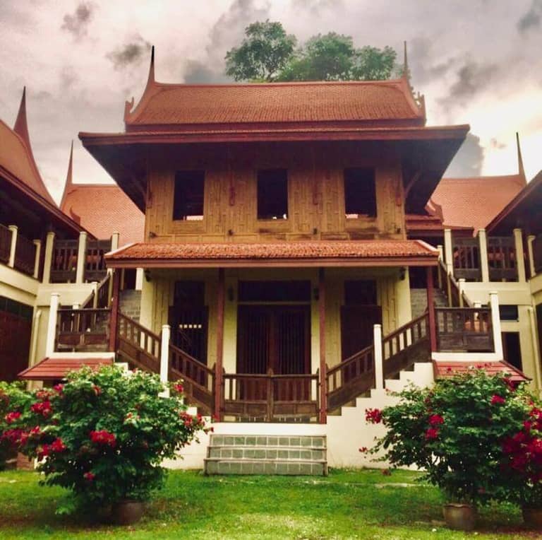 A traditional Thai wooden house with steep gabled roofs in Luang Chumni Village, Ayutthaya, surrounded by flowering bushes and greenery under a cloudy sky.