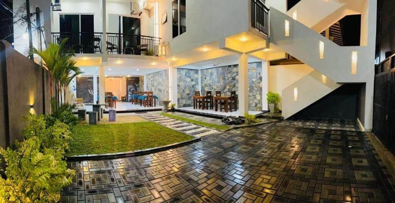Modern two-story Funwhales House in Negombo features large windows, outdoor dining areas, stone walls, and a well-lit driveway, all surrounded by lush green plants and a neatly manicured lawn at night.