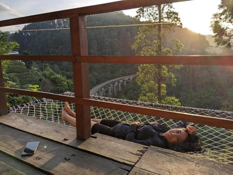 A person lies on a hammock next to a wooden railing at Barn Hills Ella, overlooking a lush green landscape with a curving railroad bridge and trees at sunset. A smartphone rests on the wooden floor nearby.