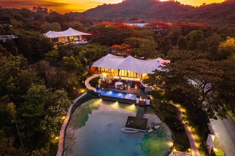 Aerial view of a luxury villa with a pool at Te Mata Glamping, surrounded by dense trees and Khao Yai hills at sunset, with warm lighting illuminating the property for an unforgettable glamping experience.