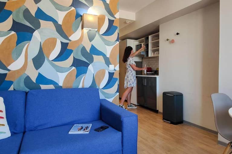 In a stylish Nice aparthotel by AMMI, a woman in a floral dress reaches into a kitchen cabinet, surrounded by a modern apartment with a blue couch and patterned wall.