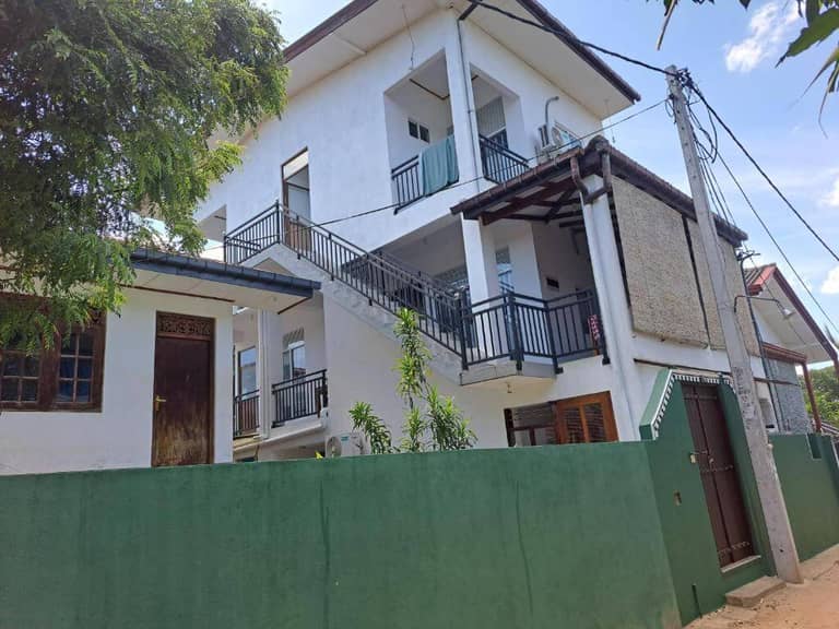 Two-story white building with balconies and exterior stairs, surrounded by a green wall and gated entrance, under a partly cloudy sky—welcome to B & B CHARM Dikwella.