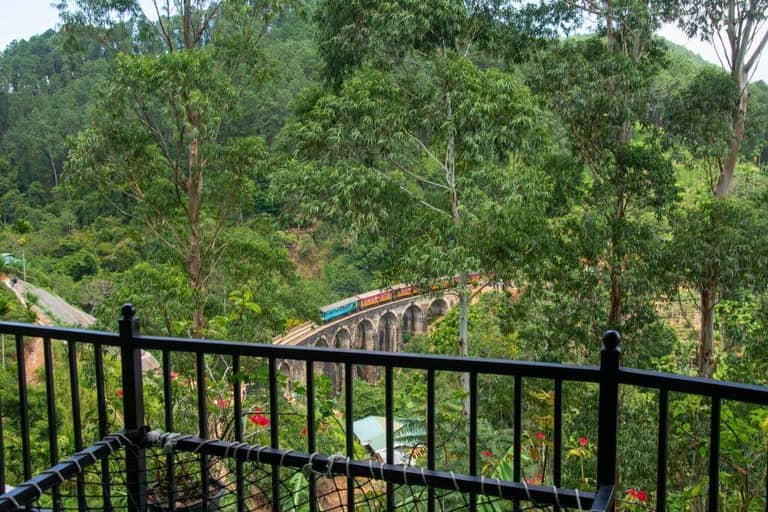 A train crosses the iconic 9 Arch viaduct bridge near Ella, winding through a lush forest and framed by a black metal balcony railing in the foreground, offering a stunning view.