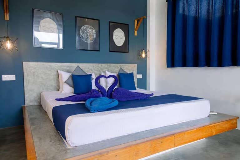 A modern bedroom at Haven Beach, Hiriketiya, featuring a double bed with blue and white bedding, towel swans on the bed, three wall art pieces above the headboard, and a window dressed in dark blue curtains.