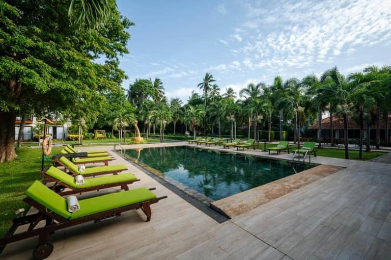 Rectangular outdoor swimming pool surrounded by green lounge chairs, palm trees, and a tiled pool deck under a blue sky.