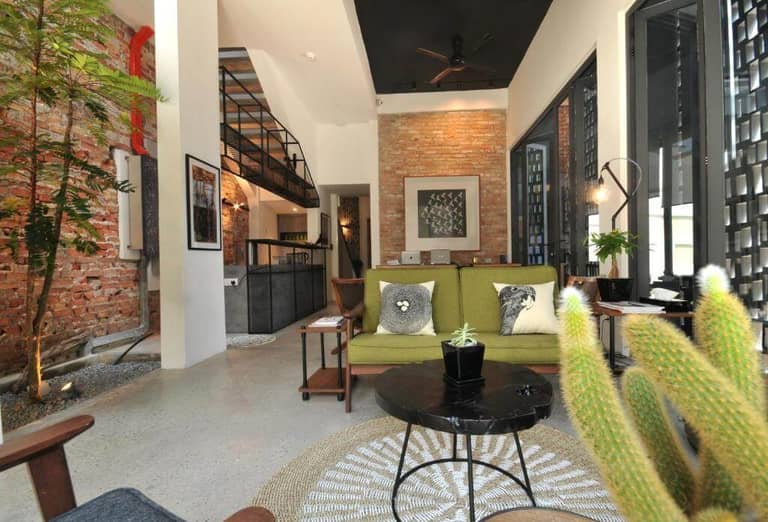 Modern living room at Nest House Melaka with exposed brick walls, green sofa adorned with animal pillows, black coffee table, lush indoor plants, and a stylish loft area featuring a metal staircase.