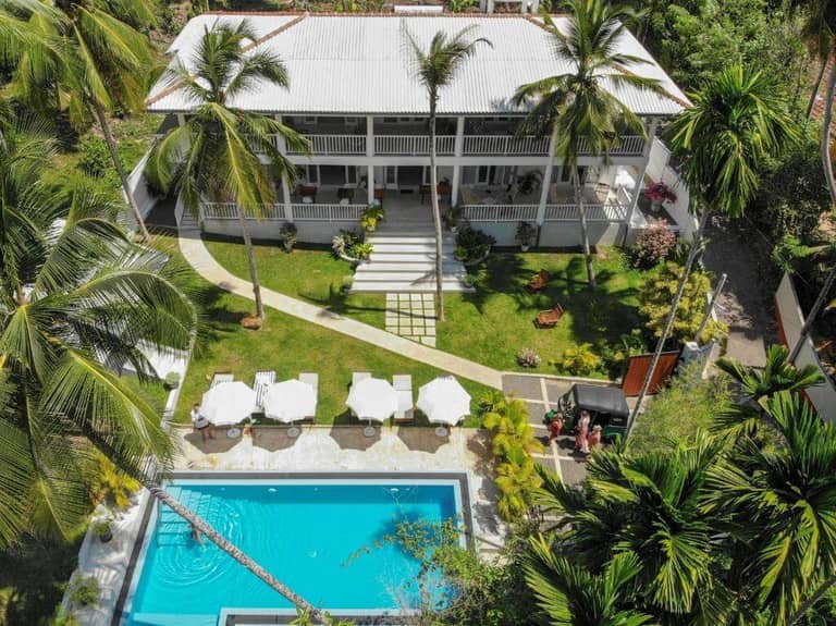 Aerial view of Plantation House, a two-story white building with a veranda in Hikkaduwa, surrounded by palm trees, a lawn, sun loungers, umbrellas, and a rectangular swimming pool.