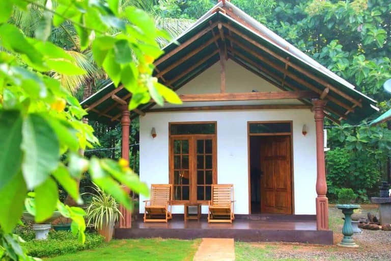 A small white cottage with a wooden porch, two chairs, and a wooden door, surrounded by green plants and trees at Green View Udawalawe.