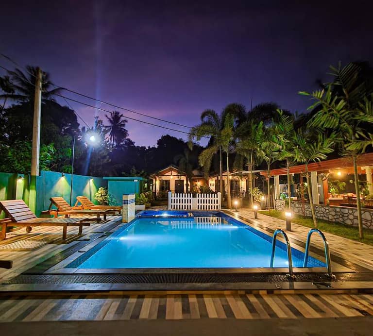 The outdoor swimming pool at Max Villa Udawalawe is illuminated at night, surrounded by lounge chairs, swaying palm trees, and lit buildings in the background.
