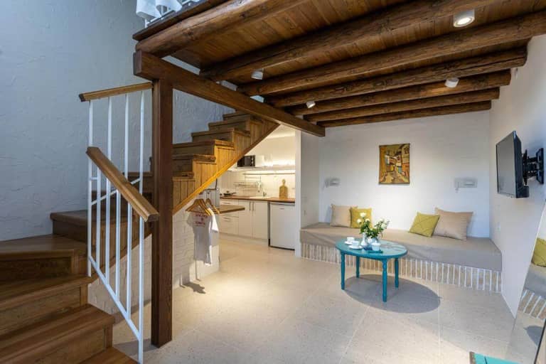 Modern living space by Khujra Studios with exposed wooden beams, a staircase, small kitchen area, wall-mounted TV, and a cozy seating area with a round table and decorative cushions inspired by Bukhara.