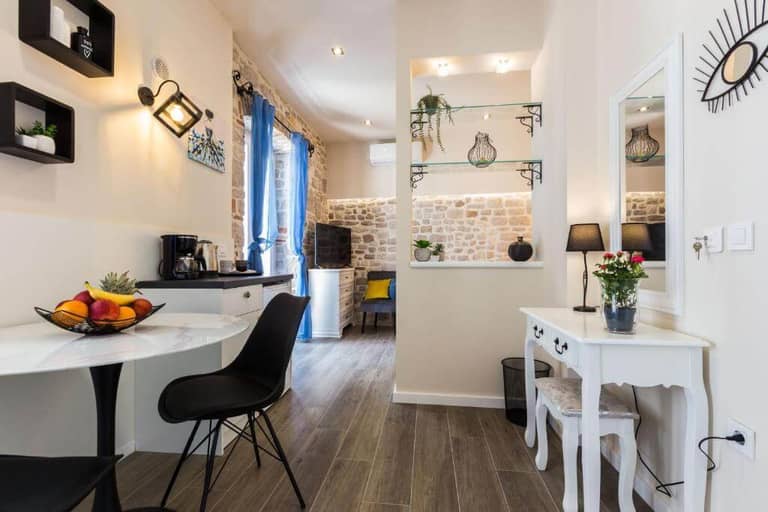 Modern studio apartment in Zadar with stone accent wall, small dining area, and desk. Features decorative shelves, blue curtains, and neutral-toned furnishings—perfect for a cozy stay at Angie Rooms.