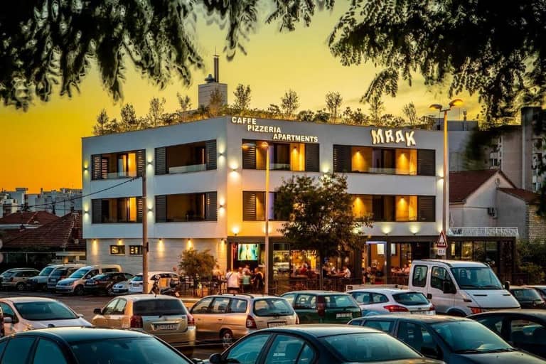 A modern three-story building labeled MRAK with a cafe, pizzeria, and apartments is shown at sunset, with parked cars in front and people sitting on the outdoor terrace.