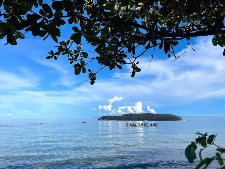A view of Sumilon Island across calm blue water, with leafy branches framing the top of the image under a blue sky with clouds, showcases the serene beauty near Bangcogon Resort in Oslob.