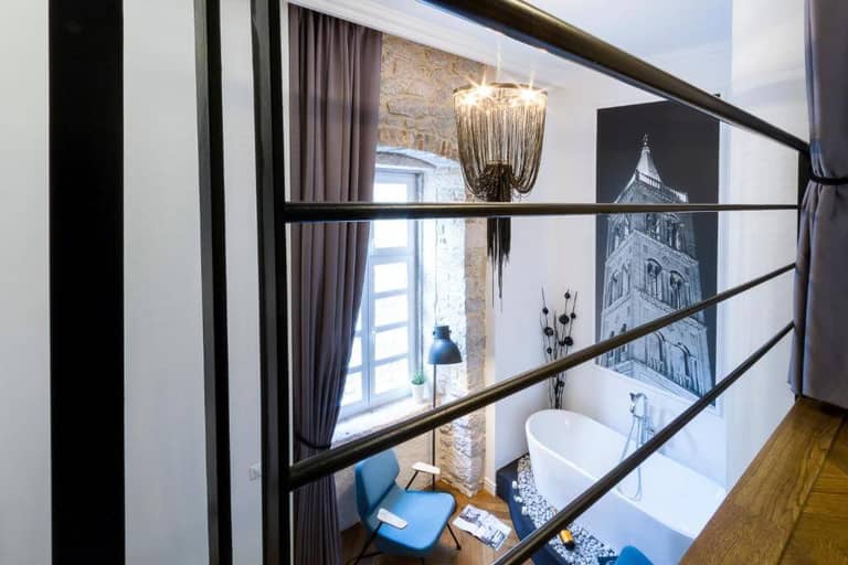 Modern loft interior at DeZign Apartments featuring a freestanding bathtub, blue chairs, exposed brick wall, large window, and chandelier—all viewed from an upper level behind black railing bars in Zadar.