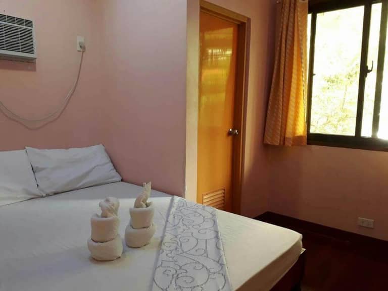 A neatly made bed with white linens and towel sculptures sits next to a window with yellow curtains and a closed door in a small room with peach-colored walls at David's Guest House in El Nido.