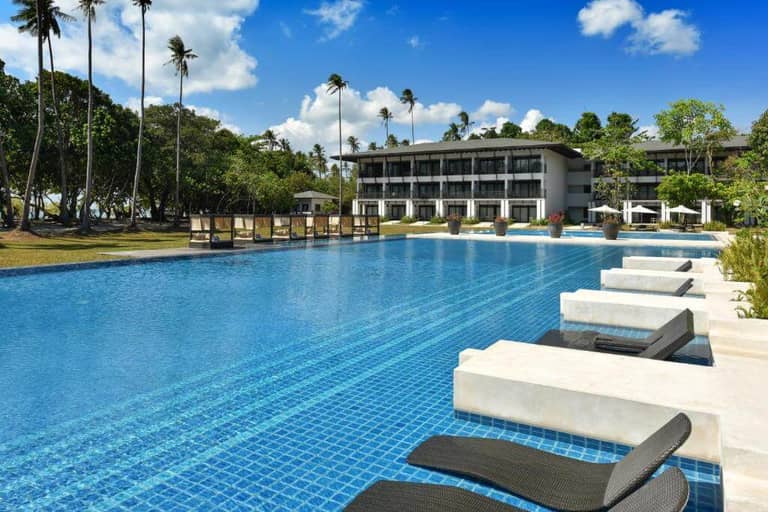 Large outdoor swimming pool with black lounge chairs, white platforms, and the multi-story Seda Lio hotel in the background, surrounded by palm trees under a blue El Nido sky.