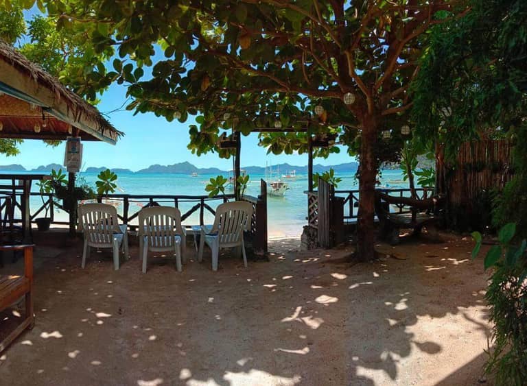 Shaded outdoor seating area with plastic chairs at Corong Beach Resort overlooks a beach, clear blue water, sailboats, and distant islands under a bright El Nido sky.