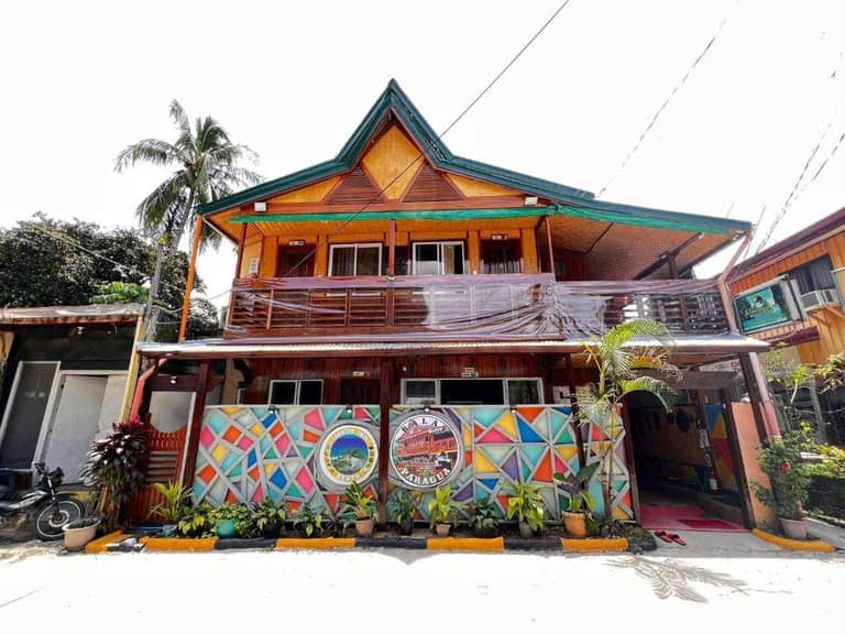A two-story wooden building in El Nido with a geometric mural and plants in front, featuring a balcony and triangular roof, a palm tree on the left side, and inspired by Balay Paragua’s distinctive local charm.