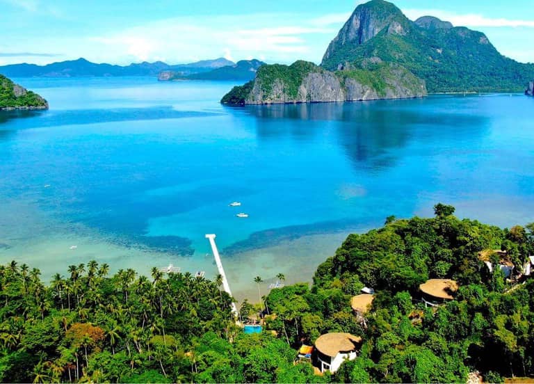 Aerial view of a tropical coastline in El Nido with clear blue water, lush green trees, a small pier, and distant rocky islands under a sunny sky—an idyllic escape near LIHIM RESORTS.