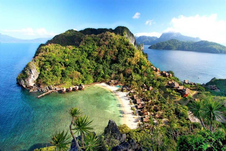 Aerial view of El Nido’s tropical island with lush green hills, a sandy beach, clear blue water, and Cauayan Island Resort’s wooden huts along the shoreline.
