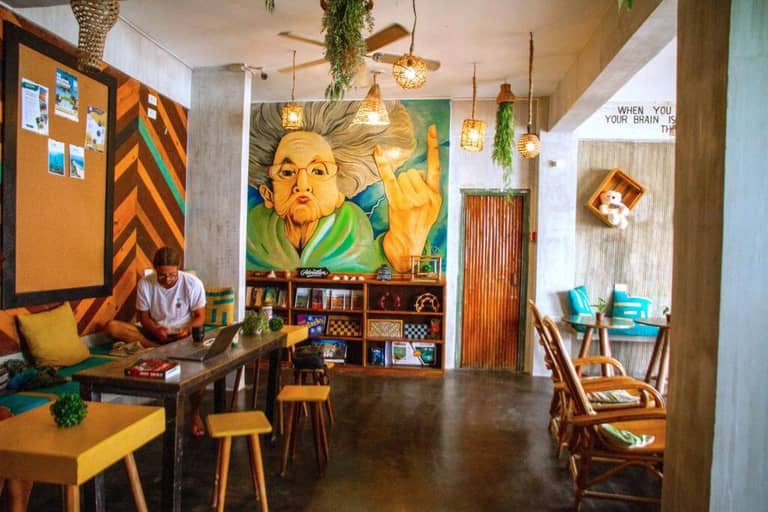A cozy, colorful cafe interior at Happiness Hostel in El Nido, with wooden furniture, wall art of an elderly woman, potted plants, and a person using a laptop at a table.
