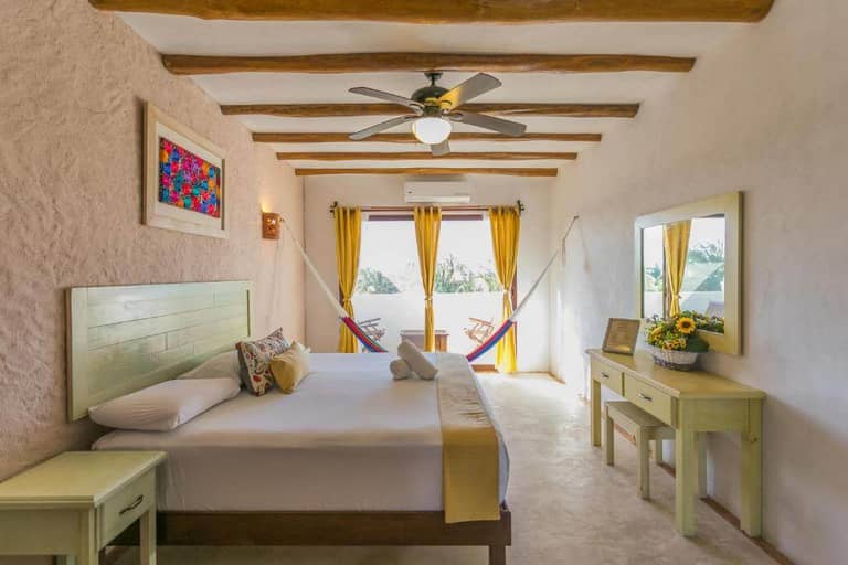 A bright bedroom at Villas Margaritas, Holbox, with a double bed, ceiling fan, wooden beams, a desk with a mirror, two hammocks by a window, and yellow curtains.