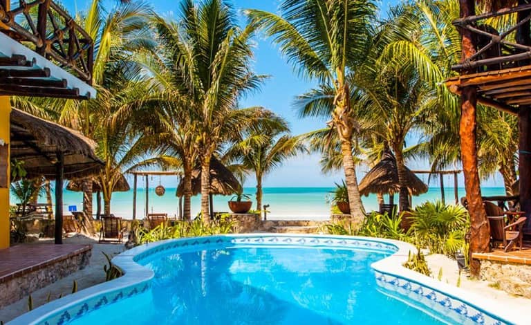 A swimming pool surrounded by palm trees at Holbox Dream, a beachfront hotel, with a stunning view of the beach and ocean in the background on a sunny day.