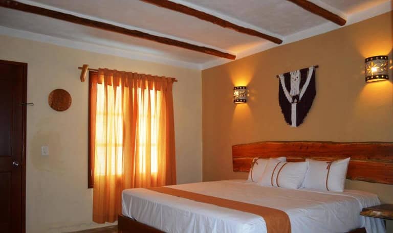 A cozy bedroom at Casa San Juan in Valladolid features a large bed with a wooden headboard, orange and white bedding, sunlit curtains, and warm-toned walls adorned with rustic decor.