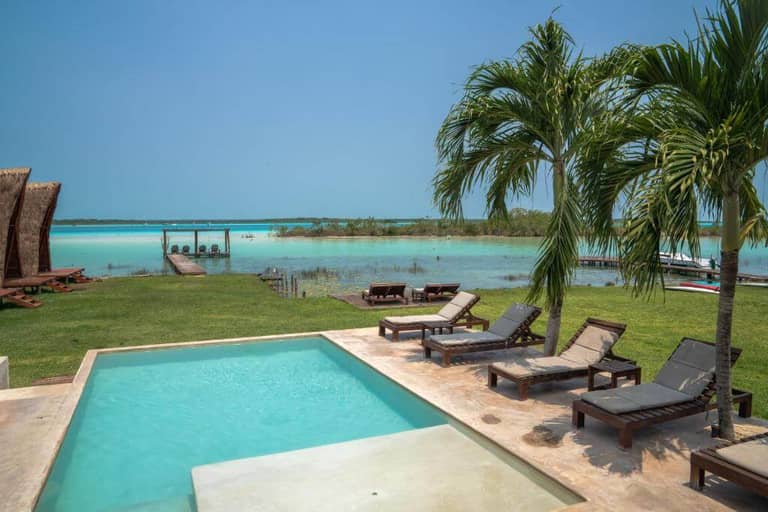 A small pool with lounge chairs is set on a patio at CasaBakal, overlooking a grassy area, palm trees, and Bacalar’s calm turquoise lake with a wooden dock in the distance.