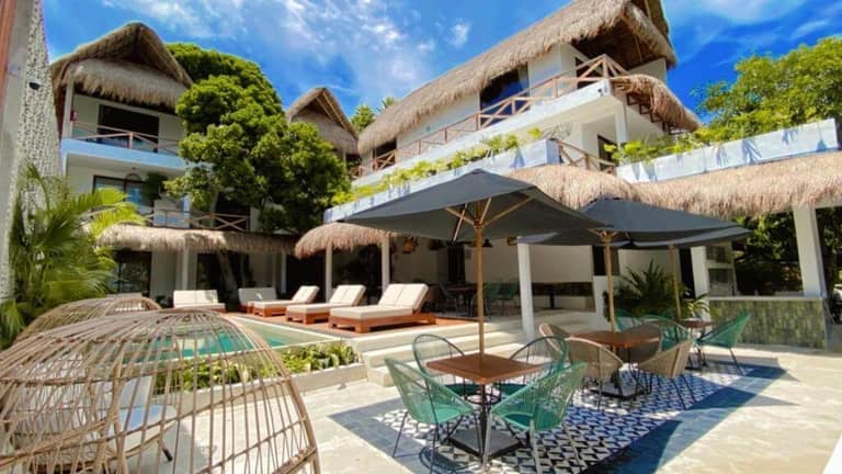 A modern tropical Bacalar hotel features thatched roofs, outdoor lounge chairs by a pool, shaded dining tables, and lush greenery under a blue sky.