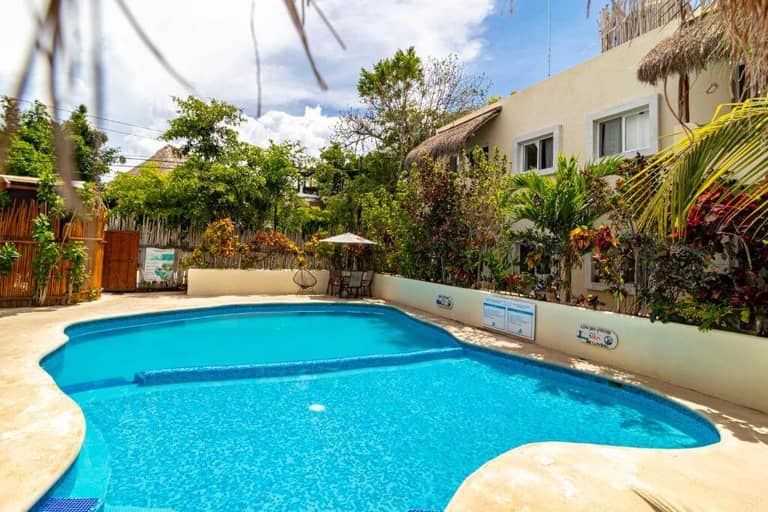 Outdoor swimming pool surrounded by tropical plants and a light-colored building with windows; lounge chairs and a small table are visible in the background at Hotel Oasis Bacalar, a welcoming Bacalar hotel.