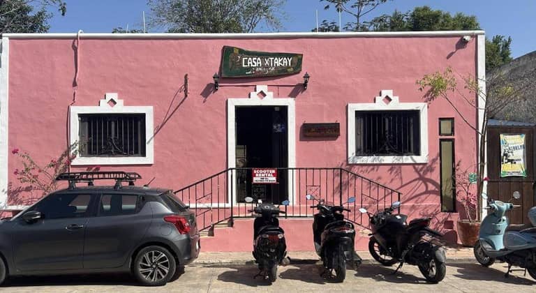 A pink building labeled Casa Kitxkay stands in Valladolid with two windows, a central door, and a Scooter Rental sign by the entrance. Three scooters and a car are parked out front of this welcoming hostal.