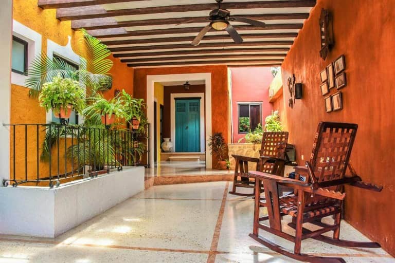 Charming covered patio at Casa Tia Micha in Valladolid, featuring orange walls, potted plants, wooden rocking chairs, and a blue door at the end of the walkway. Ceiling fan and framed decor add cozy touches.