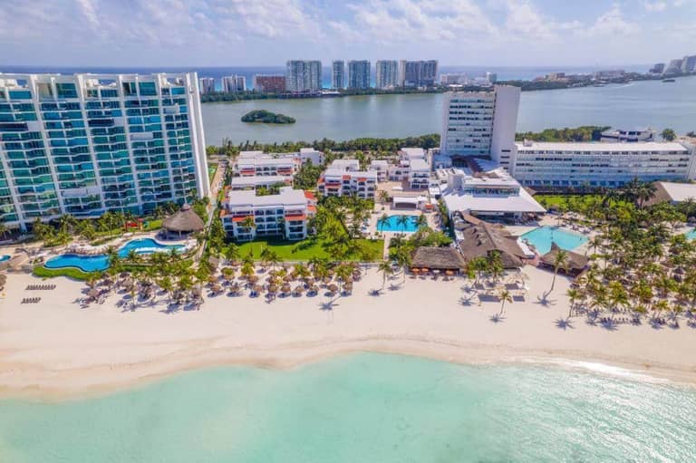 Aerial view of Beachscape Kin Ha Cancun, featuring pools, palm trees, and lounge chairs along white sand and turquoise water; stylish Villas & Suites overlook the lagoon with city buildings in the background.