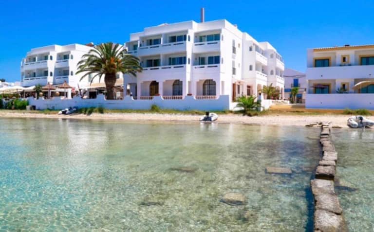 White waterfront hotel buildings with balconies, including Sabina Suites, are situated next to Formentera’s clear, shallow sea, with palm trees and boats visible on a sunny day.