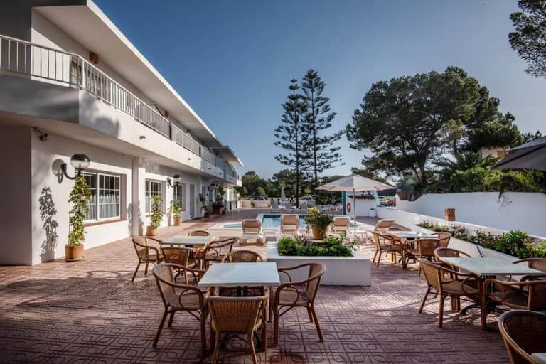 Outdoor dining area with wicker chairs and white tables on a tiled patio at Hostal Es Pi, overlooking a pool and surrounded by trees and a two-story white building in beautiful Formentera.
