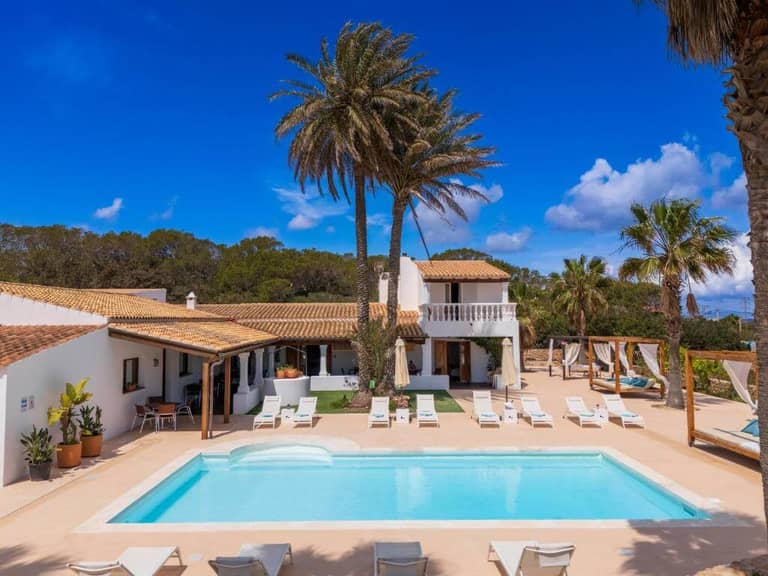 A large villa in Es Pas Formentera with a swimming pool, surrounded by lounge chairs, palm trees, and clear blue sky, featuring a shaded terrace and white exterior walls.