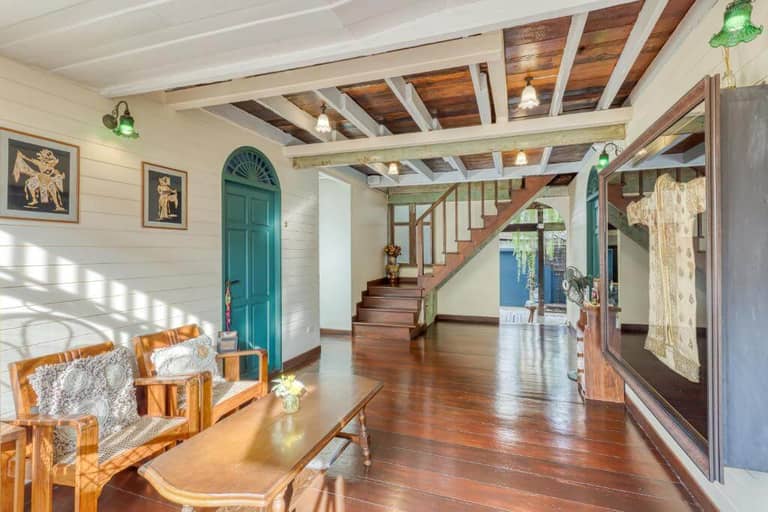 A bright room at Priya Boutique House in Bangkok features wooden floors, exposed beam ceiling, a staircase, green door, cozy sofa set, coffee table, and framed art on the wall.