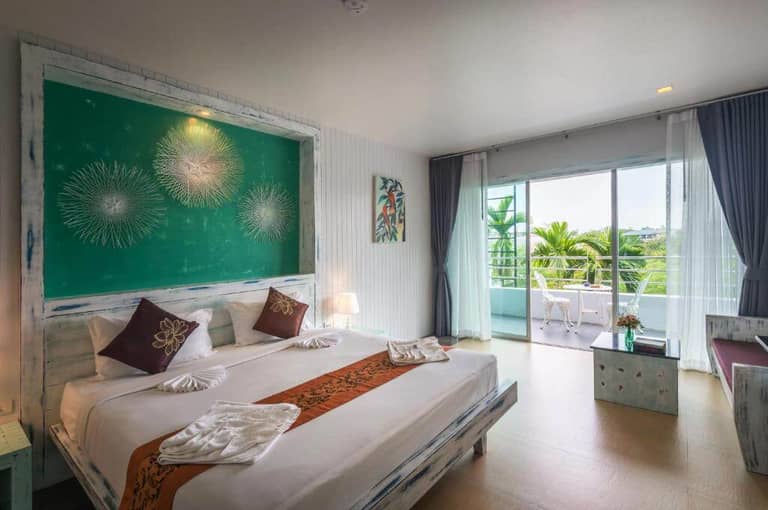 Modern hotel room at Baan Ban Dalay in Krabi, featuring a large bed, green accent wall, balcony with outdoor seating, and natural light streaming through sliding glass doors.