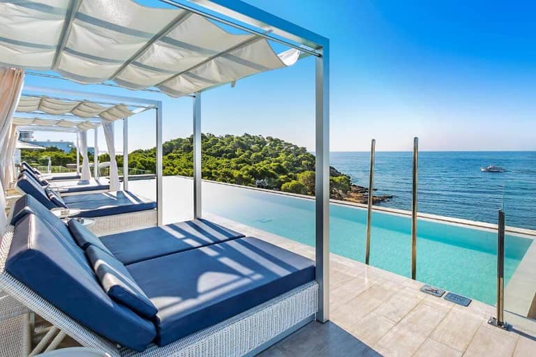 Poolside cabanas with blue cushions beside an infinity pool overlook the ocean and green hillside at Catalonia Royal Ses Estaques, an adults only hotel in Ibiza under a clear blue sky.