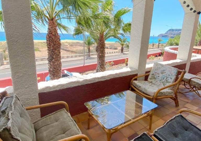 A covered patio at this Destination Hotel features wicker chairs and a glass table, overlooking Porto Santo’s coastal road, palm trees, and the ocean under a clear blue sky.
