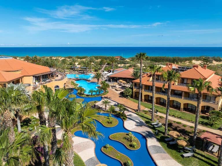 Aerial view of Pestana Porto Santo beachfront resort with terracotta-roofed buildings, winding pools, palm trees, and gardens, overlooking the ocean under a clear blue sky.